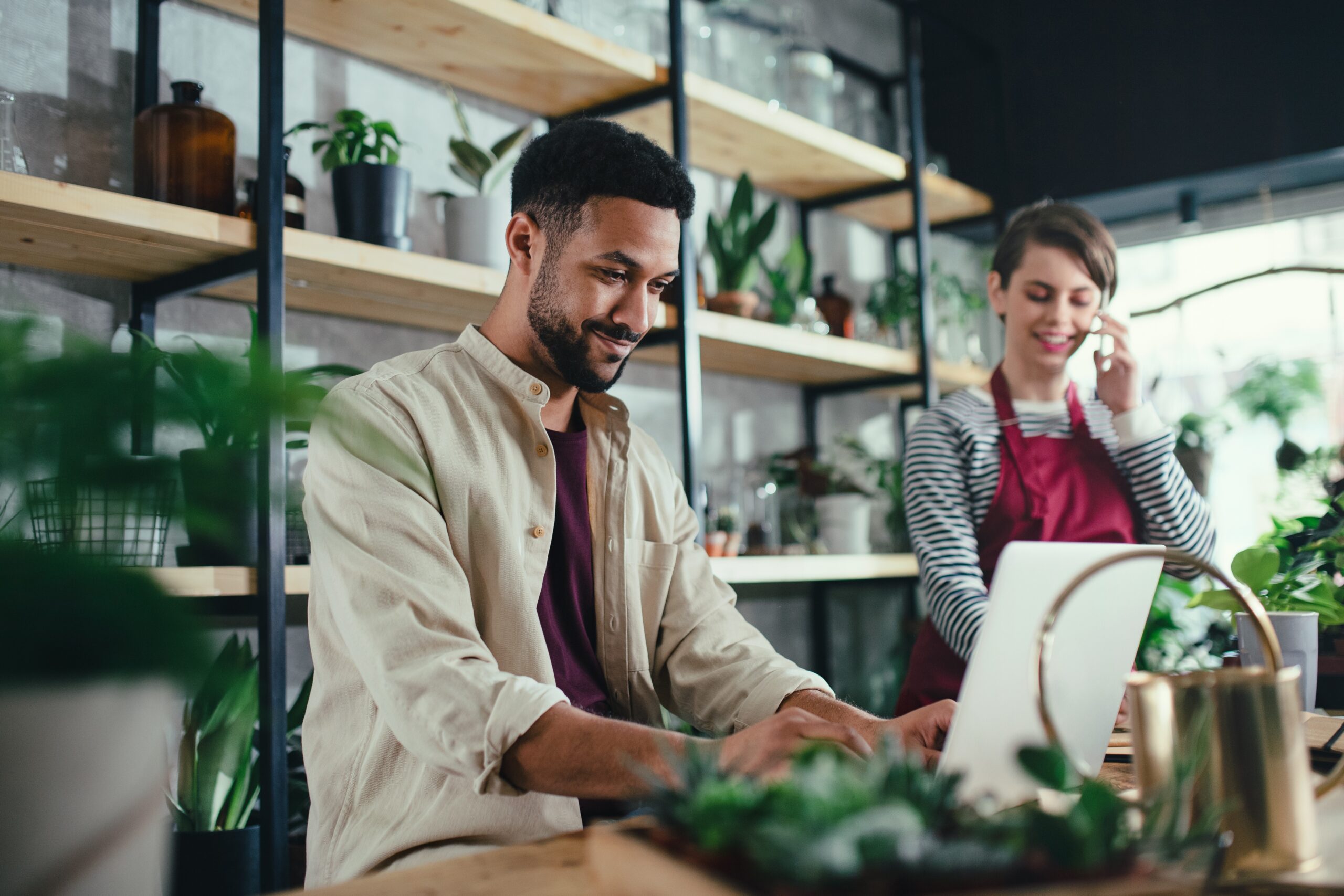 A shop owner at a laptop while a colleague takes a call in a plant-filled store
