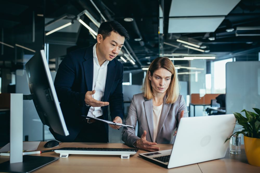 A manager standing with a clipboard talks through an invoice with a colleague at a laptop in an open-plan office
