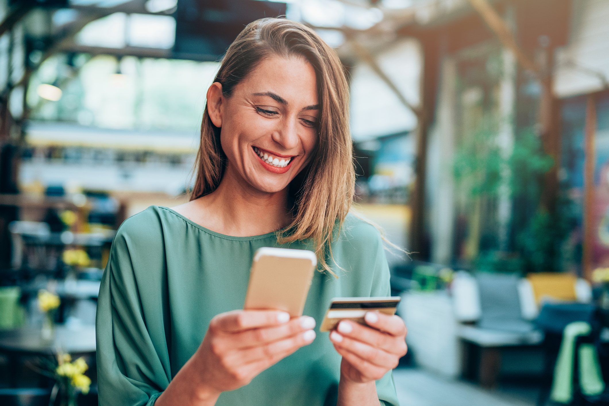 A woman in a cafe smiling at her phone while holding her bank card