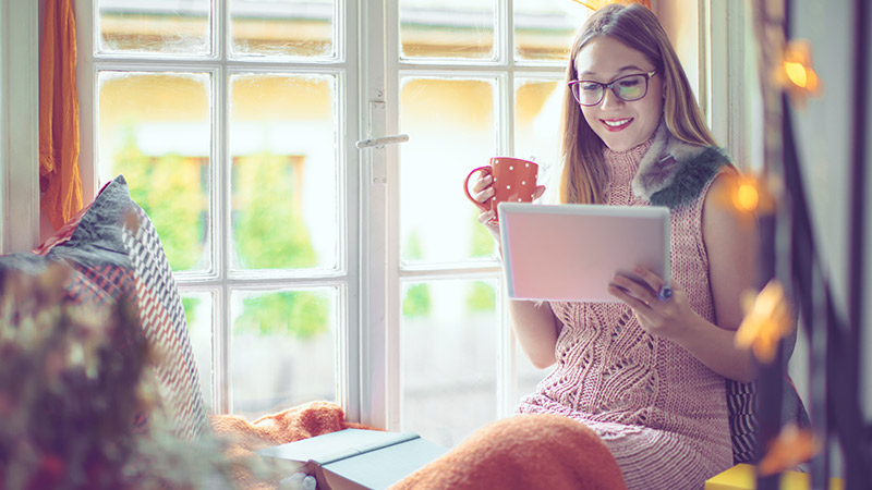 A woman on a window seat holding a mug and reading a tablet in soft daylight