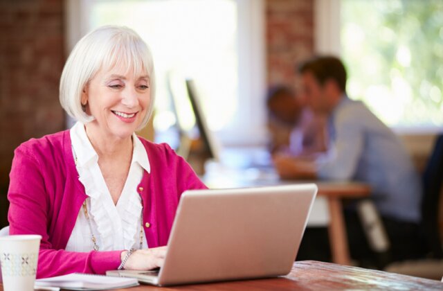 A woman with white hair smiling at her laptop in a bright, brick-walled office
