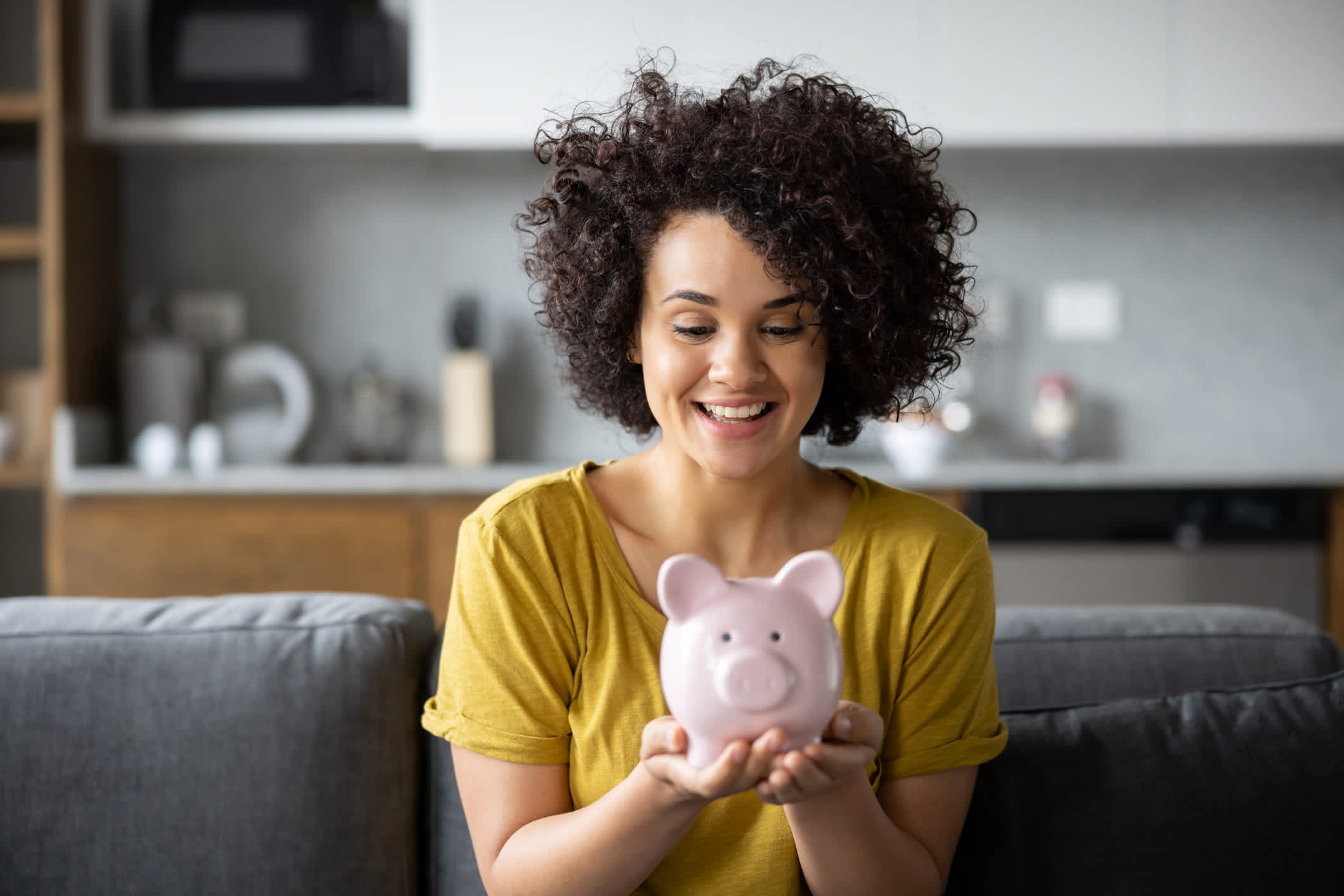 A woman sitting on a sofa, smiling at a pink piggy bank held in both hands