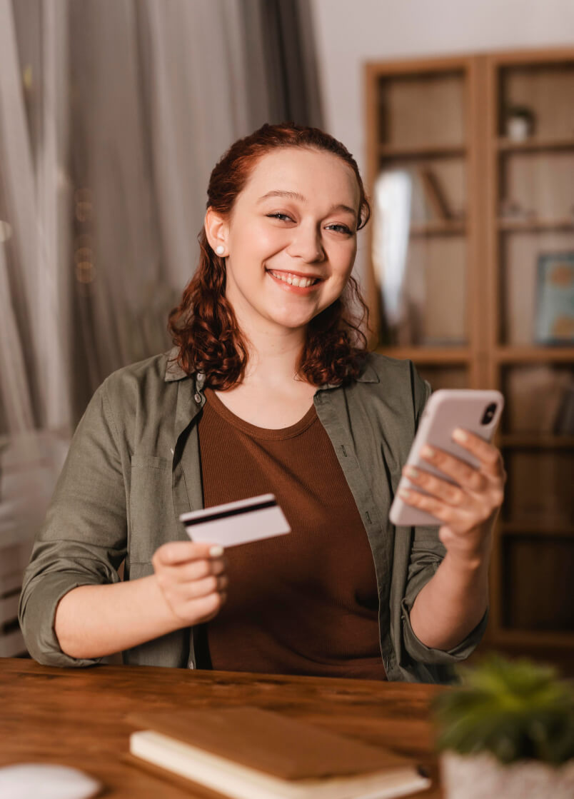 A smiling woman at a desk holding a bank card in one hand and her phone in the other
